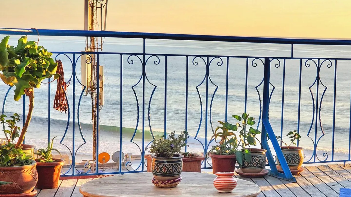 Le rooftop de Kekai Surf House au coucher du soleil, la rambarde bleue et la vague qui deroule sur la baie de Taghazout